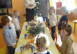 Pani dyrektor Maria Królikowska, pani Anna Tylman oraz dzieci stoją wokół nakrytego białym obrusem i poczęstunkiem stołu.